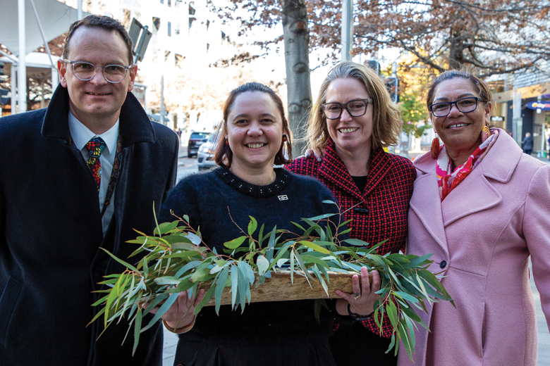 Four smiling people (one man and 4 women) stand on a city street. One person in the centre holds a wooden tray with green foliage.
