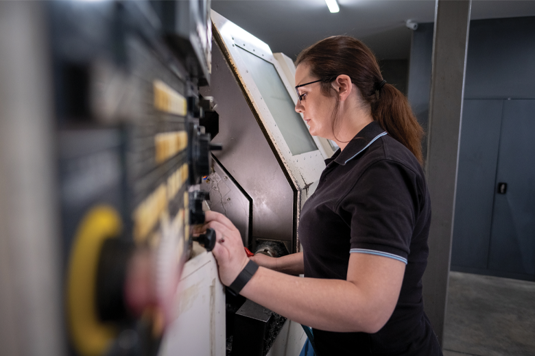 A younger woman in a black shirt is operating a machine in an industrial setting. The machine features buttons and controls. 