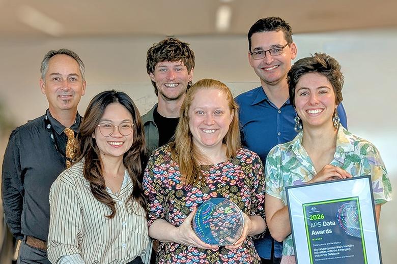 Six DISR staff standing in an office with white walls and grey carpet. The people are positioned across two rows, smiling and professionally dressed. The woman in the centre of the front row is holding a circular award, and the woman to the right of her is holding a framed certificate with the words ‘2026 APS Data Award’ visible. 