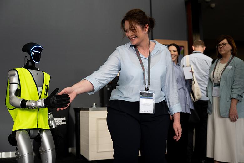 In a grey room, a humanoid G1 robot stands on the left-hand side of the image, wearing a yellow high visibility vest and a black glove on one hand. On the right-hand side of the image is Casey Martone from the National AI Centre, wearing a blue shirt with a lanyard around her neck, smiling down at the robot. The robot has its gloved hand out-stretched and is shaking Casey’s hand. Three people are standing in the background also wearing lanyards. 
