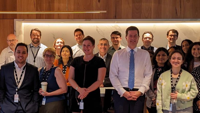 A large group of DISR staff stand gathered in an office break room with wooden flooring, smiling. The staff are dressed professionally and mostly wearing lanyards with ID cards attached. Deputy Secretary Robert Jeremenko stands in the front row, to the right of the image, wearing a white shirt and a blue tie.