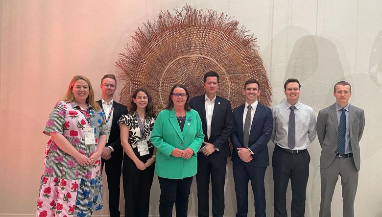 A group of eight DISR representatives, smiling and dressed in professional clothing, stand in a room against a white wall. The Hon Madeleine King MP, Minister for Resources and Minister for Northern Australia, stands in the centre of the image, wearing a green jacket. Standing to the right of her is Deputy Secretary Robert Jeremenko, who is dressed in a black suit. Three DISR representatives stand to the left of the Minister. Three DISR representatives stand to the right of Robert.