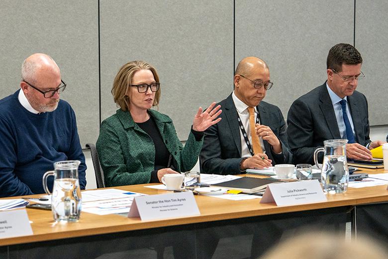 Four people sit behind a long desk in a conference room, with nameplates and papers in front of them. Senator the Hon Tim Ayres, Minister for Industry and Innovation and Minister for Science, sits at the far left wearing a dark blue jumper and reading papers on the desk in front of him. To his right, Acting Secretary Julia Pickworth, wearing a green jacket, speaks while gesturing with her hand to people not visible in the image. Carl Chang and Deputy Secretary Robert Jeremenko sit to her right.