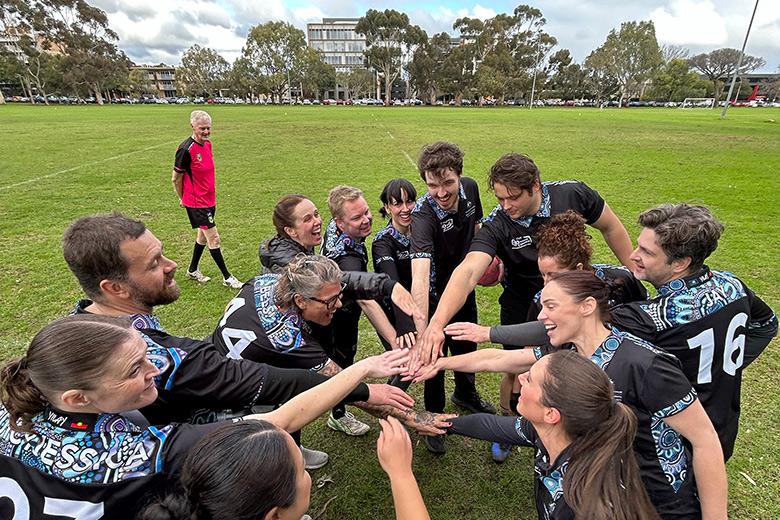 A group of DISR staff belonging to the DISR touch football team stand in a circle on an oval. They are wearing black shirts with a blue artwork celebrating NAIDOC Week. The players are standing in circle with their hands joining in the centre for a hand stack.