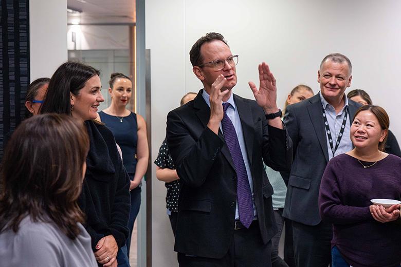 Deputy Secretary Neal Mason addressing a group of professionally dressed DISR staff in an open white workspace. Neal is wearing a black suit with a purple tie and is gesturing with his hands. Staff are gathered around smiling and listening.