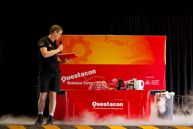 A man on a stage, dressed in a black shirt and shorts, speaks into a microphone and gestures towards a red and orange Questacon display. Smoke is coming from a bin to the right of the display, indicating that he is presenting a scientific experiment.