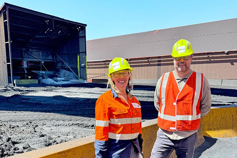 A woman and a man dressed in orange high visibility vests and yellow hard hats, standing outside the Whyalla steelworks site in South Australia. Behind them is a short yellow wall and an open shed. The ground is covered in grey sand. 