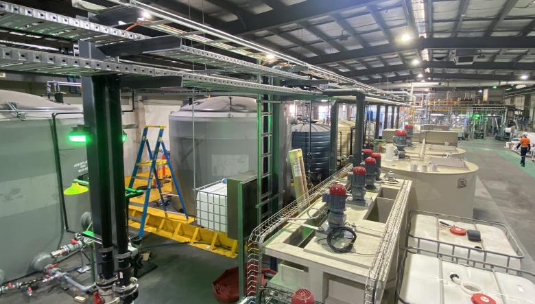 An industrial processing facility showing large storage tanks, overhead piping, pumps, control equipment, and maintenance ladders. 