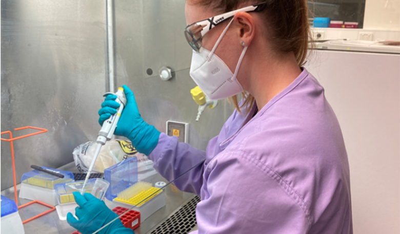 Woman in a laboratory with mask, protective glasses, gown and gloves handing a medical sample.