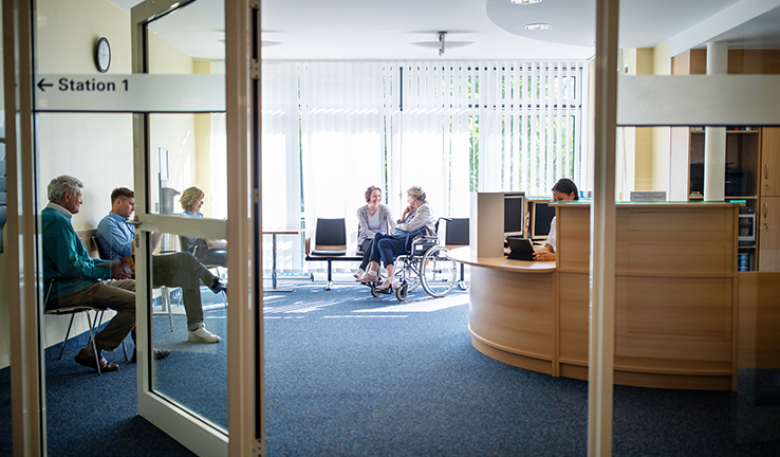 Photo of a public building entry with wide doors open and easy to get into with a wheelchair. Inside people are sitting in a waiting room and a woman is talking to a woman in a wheel chair.