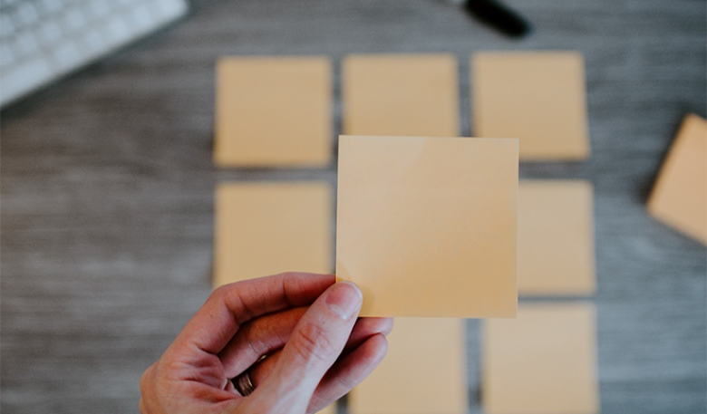 Photo of a person’s hand selecting a blank cream post it note from set of 9 on their desk. Photo of a person’s hand selecting a blank cream post it note from set of 9 on their desk.