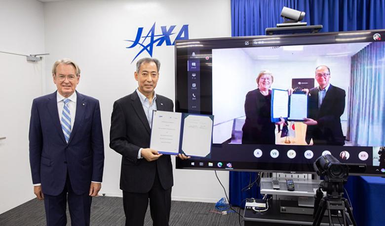Two men standing beside a tv screen, with two people featured on the screen