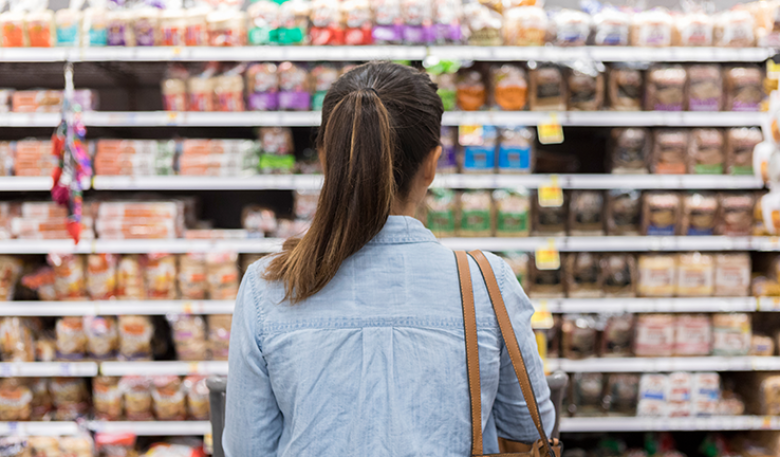 Photo of a woman in a supermarket with her shopping cart looking at products on a row of shelves.