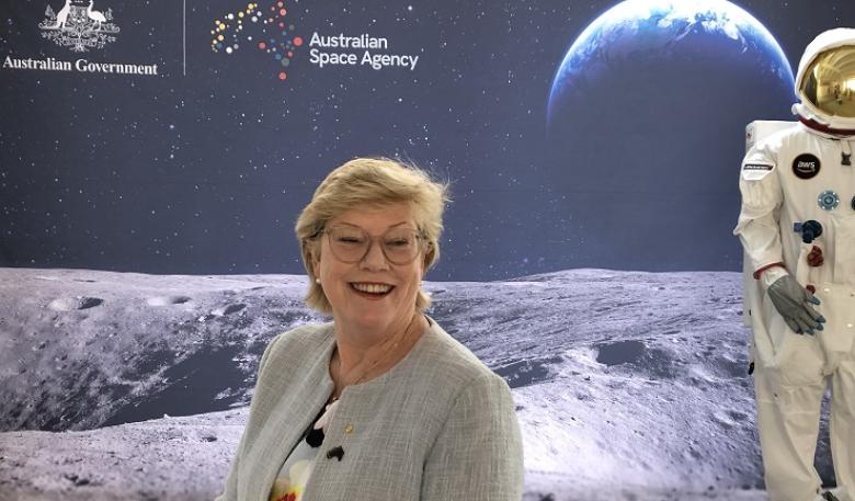 An upper body photo of a smiling woman with short blonde hair and glasses, wearing business attire. A space-related display is set up in the background.