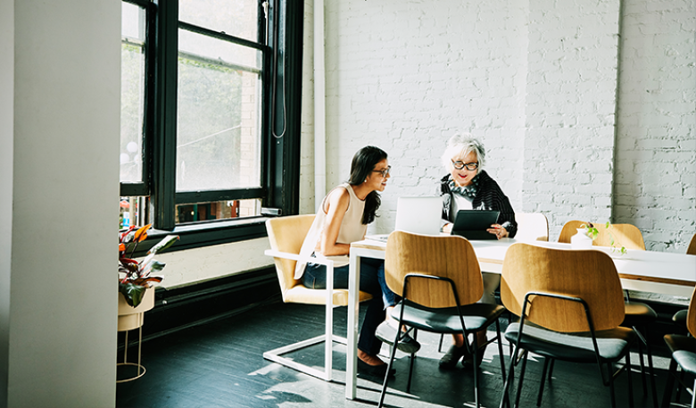 Photo of two women sitting at a table looking at a laptop and tablet