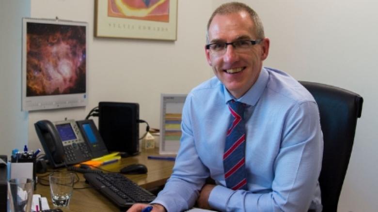 Project Director David Luchetti sitting in an office, smiling and wearing glasses, a blue shirt and striped blue and red tie. 