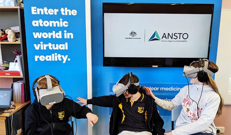 Three school children wear virtual reality helmets in front of an ANSTO display.