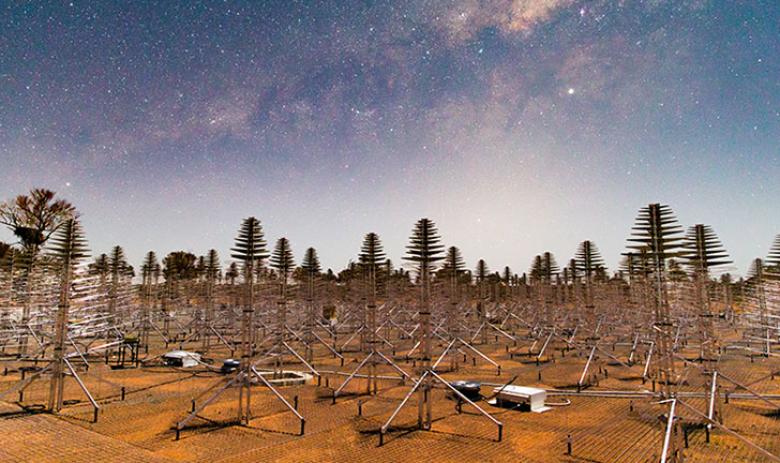 Wide angle image of metal Christmas tree-like antennas on red soil with the stars of the Milky Way overhead.