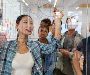 A diverse group of business people watch a female colleague draw business growth projections on a clear window.
