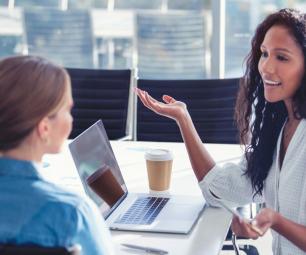 Two businesswomen have a conversation at a table with their laptops and coffee.