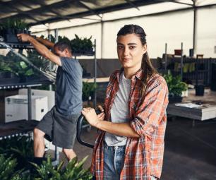 A woman in a small warehouse leans against some equipment. A man behind her lifts a tray of plants off a shelf.