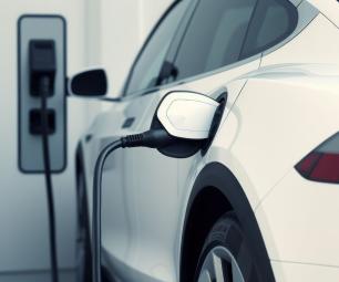 Photo of the back end of a white electric vehicle with a charging cable plugged in. The electric vehicle supply equipment (EVSE) is in the background.