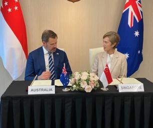 Minister Husic and Minister Teo, sitting at a desk signing the MoU. They are surrounded by flags of Australia and Singapore. 
