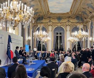 Photo of the AI Action Summit stage located in the Grand Palais. Several political figures are sitting down and talking to a room filled with people. 