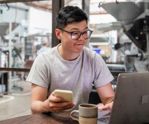 A young man on his laptop with a phone handy. He's sitting in a tech lab with an office behind him.