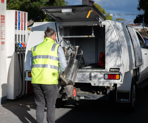 A field officer wearing a high‑visibility vest unloads fuel‑testing equipment from the back of a utility vehicle at a service station.  
