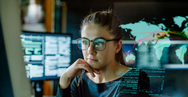 Photo of woman working in front of computer monitors in an office. In front of her, there is a see-through panel displaying a map of the world and other data.