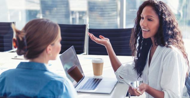 Two businesswomen have a conversation at a table with their laptops and coffee.