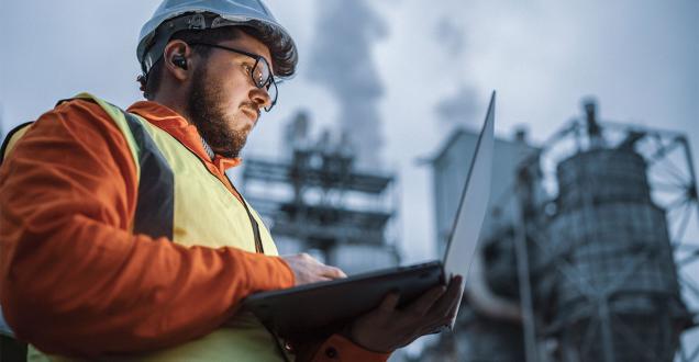 Man wearing protective gear looks at laptop while standing in front of oil and gas infrastructure.