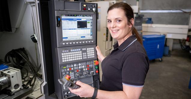 Image shows a woman in a factory. She is standing next to the control panel for a machine tool, smiling and looking towards the camera.