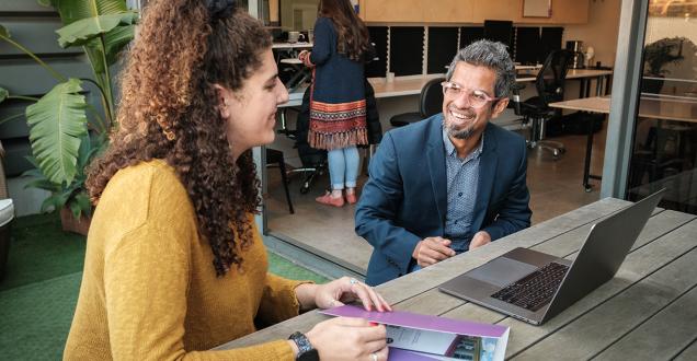 A businessman and women have a conversation at a table over a laptop. 