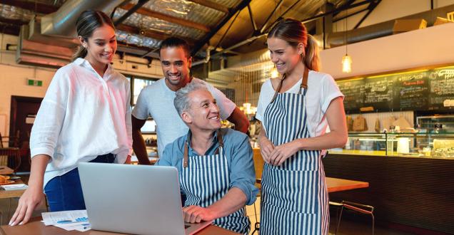 Four smiling cafe workers gather around a laptop in their place of business.