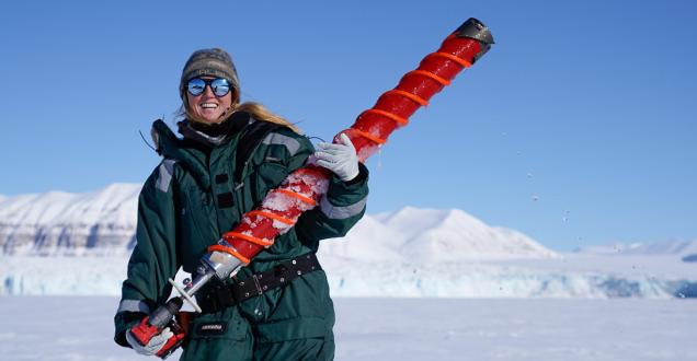 Female polar marine biologist stands in a snowy landscape holding an oversized drill bit