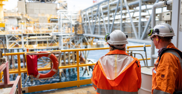 Two oil and gas workers wearing hard hats and orange gear look out from the back of a vessel onto an offshore rig.