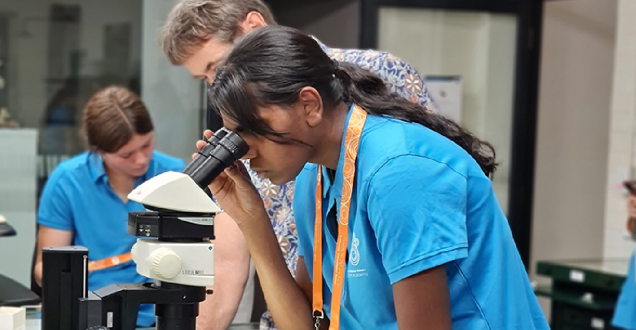 Female student looking into a microscope while a teacher looks on.
