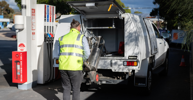 A field officer wearing a high visibility vest unloads fuel testing equipment from the back of a utility vehicle at a service station.