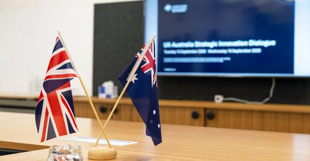 British and Australian flags on a conference room desk with a presentation screen in the background ‘UK-Australia Strategic Innovation Dialogue’. 