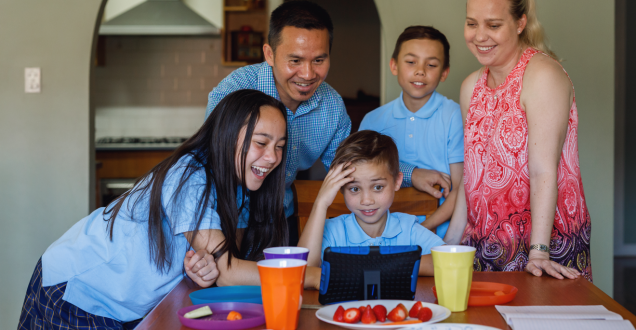 Two parents and their 3 children are smiling while they stand and sit around their dining table as they talk to family overseas using a tablet.