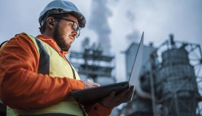 Man wearing protective gear looks at laptop while standing in front of oil and gas infrastructure.