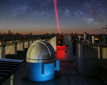 Two telescope domes on a rooftop at night, with one transmitting a red laser.