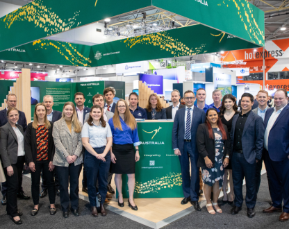 A group of people in business attire standing at a large, open plan exhibition stand at the 2023 World Mining Congress. The stand displays the Australian Government’s green and gold ‘Nation Brand’, including a kangaroo and decorative elements representing Australia’s rivers and bush tracks.