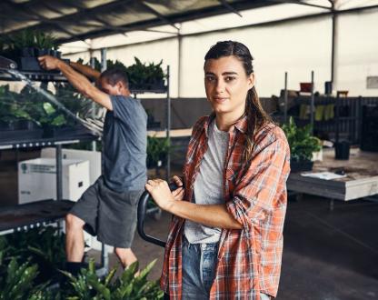 A woman in a small warehouse leans against some equipment. A man behind her lifts a tray of plants off a shelf.