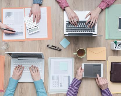 A group of people working on their computers at a desk. Their hands typing on devices that show report data. 