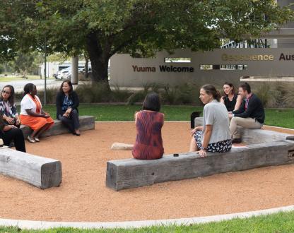 Eight female staff and a man are sitting on wooden sleepers as part of a yarning circle. In the  background, a wall has Geoscience Australia written on it, and the words 'Yuuma Welcome'.