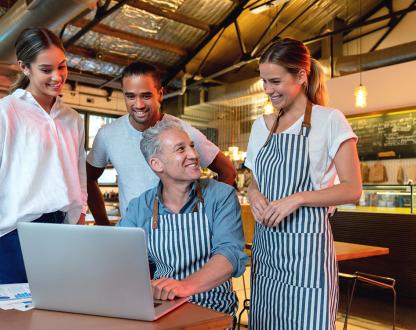 Four smiling cafe workers gather around a laptop in their place of business.