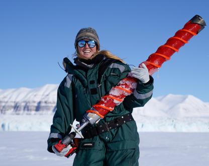 Female polar marine biologist stands in a snowy landscape holding an oversized drill bit